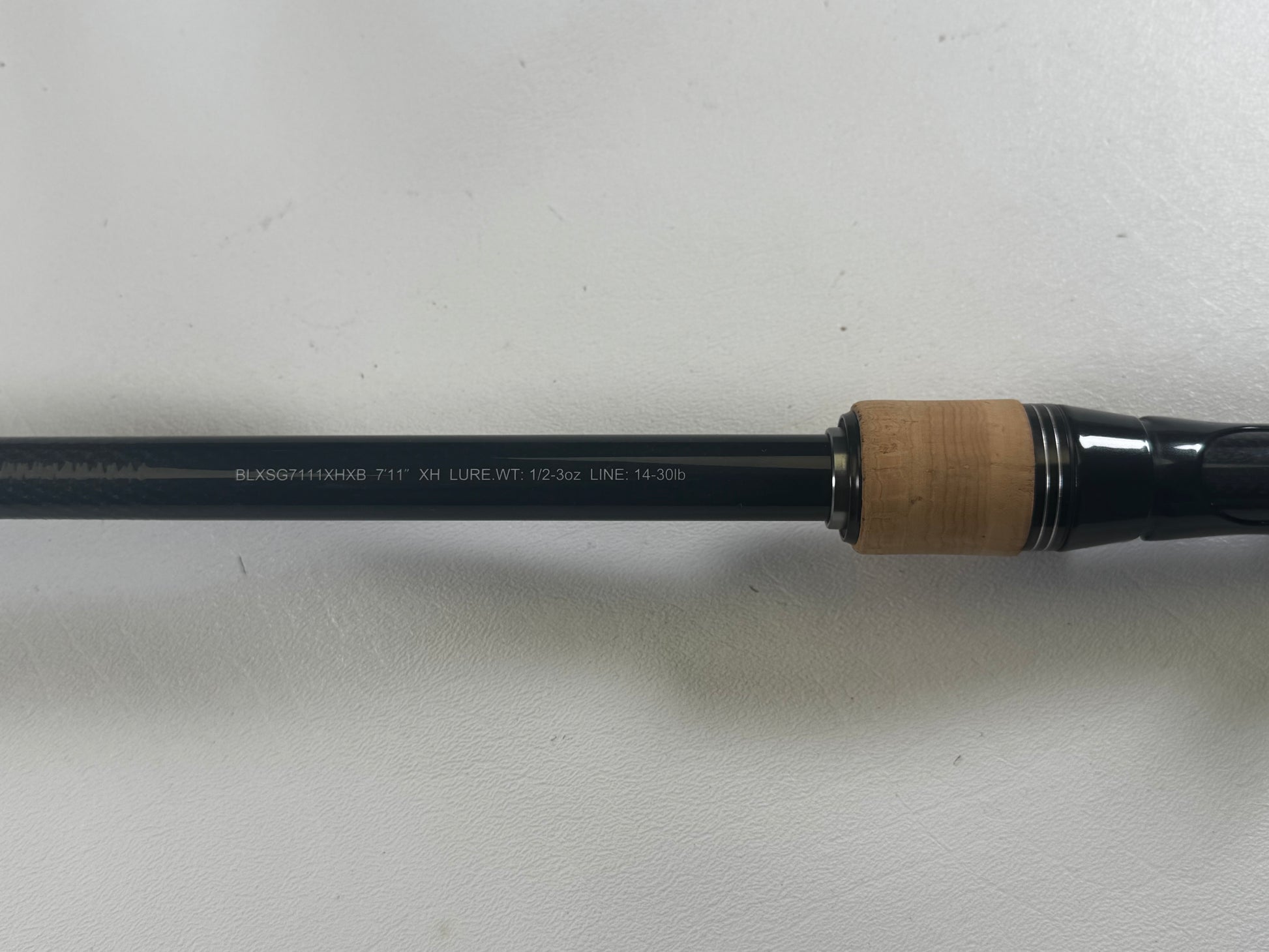 Close-up of a Daiwa BLX BLXSG7111XHXB Casting Rod showing text: "BLKSG714XHFB-7'11" XH LURE WT. 1/2-3oz LINE. 14-30lb." The cork and dark material handle is partly visible; the rod rests on a plain white surface. Excellent condition, used rod from Daiwa.