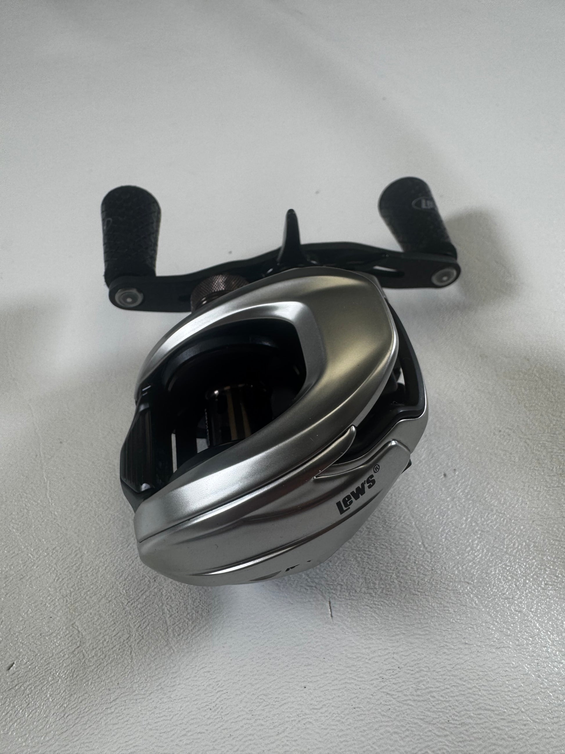A sleek, silver Lew's HyperMag TLH1XHLG2 casting reel, in excellent condition, rests on a white surface. Its black handle with textured grips is perfect for fishing adventures. Trade in your used gear and earn store credit to upgrade your fishing experience.
