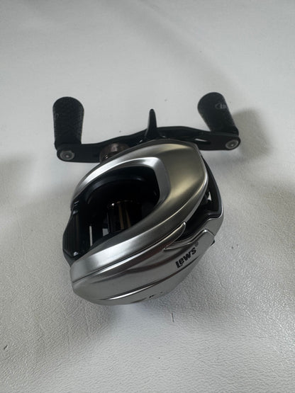 A sleek, silver Lew's HyperMag TLH1XHLG2 casting reel, in excellent condition, rests on a white surface. Its black handle with textured grips is perfect for fishing adventures. Trade in your used gear and earn store credit to upgrade your fishing experience.