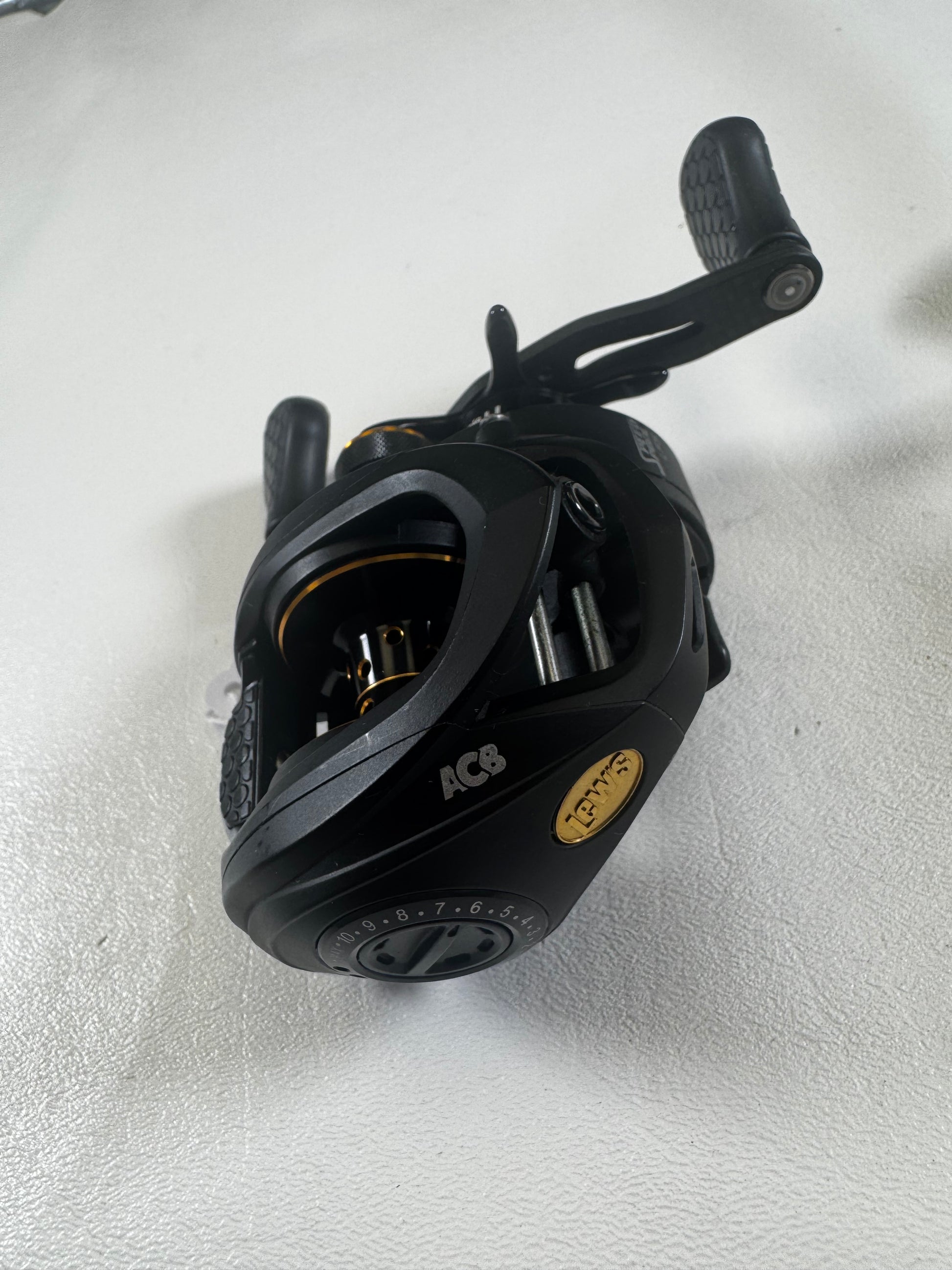 A Lew's Tournament Pro TP1SHLA 7.5:1 gear ratio baitcasting reel in excellent used condition, with visible gears and handle plus gold accents, rests on a white surface—ideal for Rod and Reel Trade-Ins or quality gear seekers.