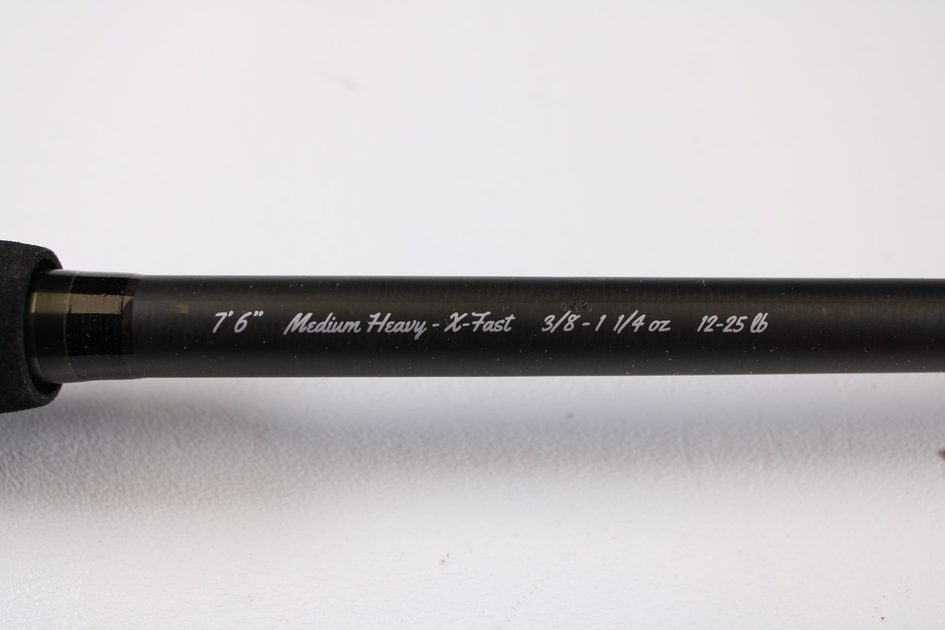 A close-up of the 13 Fishing Muse 7'6" Medium Heavy Used Casting Rod in excellent condition shows white specs: "7'6" Medium Heavy - X-Fast 3/8 - 1 1/4 oz 12-25 lb," ideal for our rod and reel trade-in program, against a light background.