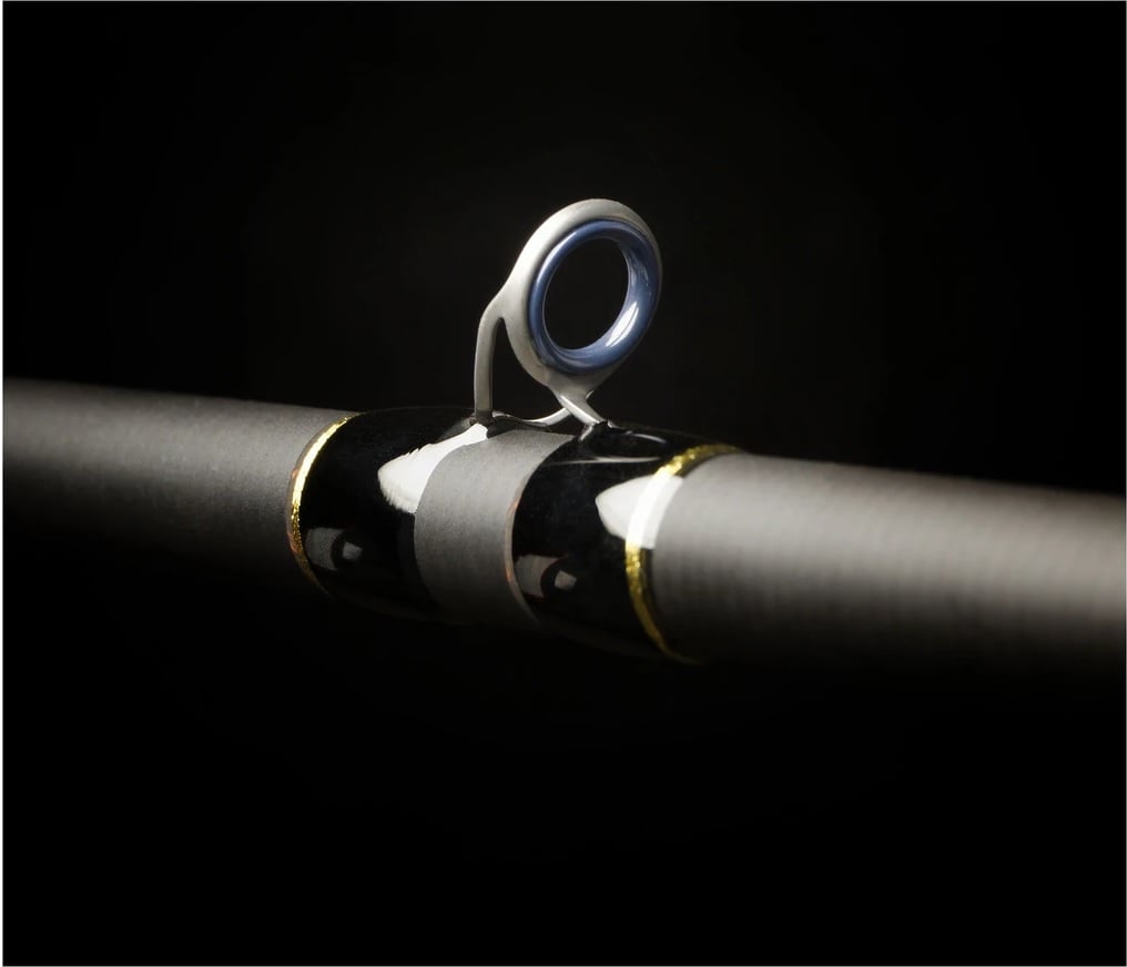 A close-up of a Falcon Cara Casting Rod Deep Runner 7'3" Medium Heavy (CC-5-173DR) guide ring, featuring smooth metallic Fuji components and black and gold wrappings, set against a dark background.