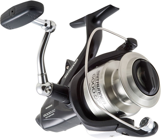 A close-up of the Shimano SHIMANO Baitrunner OC BTR6000OC Spinning Reel, highlighting its metallic silver spool, black body and handle, as well as the attached bail arm.
