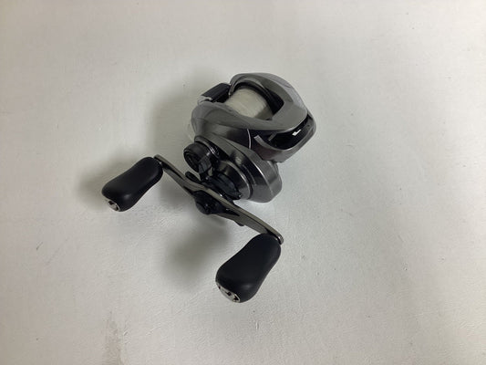 A Shimano Chronarch MGL CHMGL150HG casting reel in excellent condition, featuring a silver and black design with dual black handles, rests on a plain white surface.