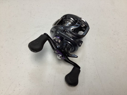 A Daiwa Tatula SV TWS TASV103XS casting reel in black and silver with two large black handles is shown on a white speckled surface. The used reel is in very good condition and made by the brand Daiwa.