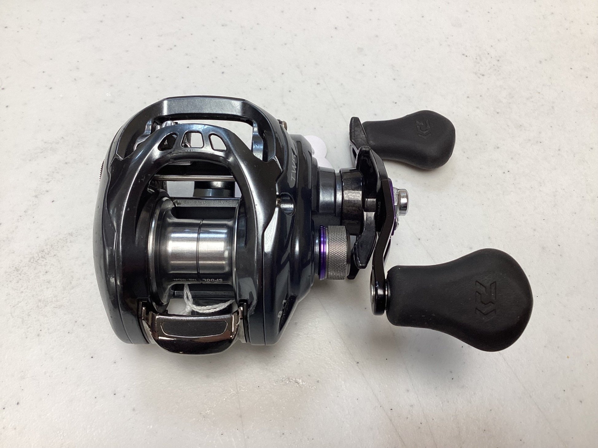 A close-up of a Daiwa Tatula SV TWS TASV103XS used casting reel in very good condition, featuring black and silver design with two black handles, shown on a white textured surface.