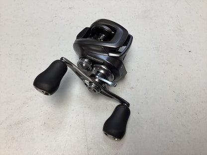 A Shimano Bantam 150XG 8.1:1 baitcasting reel, black and silver with two large handle grips, is displayed on a light-colored speckled surface.