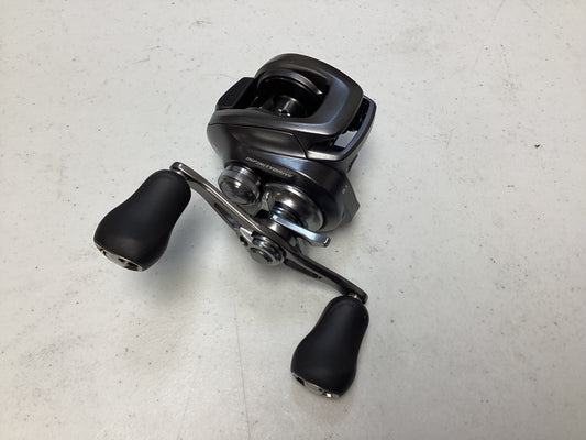 A Shimano Bantam 150XG 8.1:1 baitcasting reel, black and silver with two large handle grips, is displayed on a light-colored speckled surface.