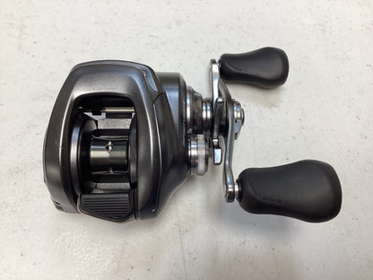 A close-up of a Shimano Bantam 150XG 8.1:1 baitcasting reel with two rubber handles, shown on a light surface. The used Shimano reel is in excellent condition, highlighting its spool and handle mechanism.