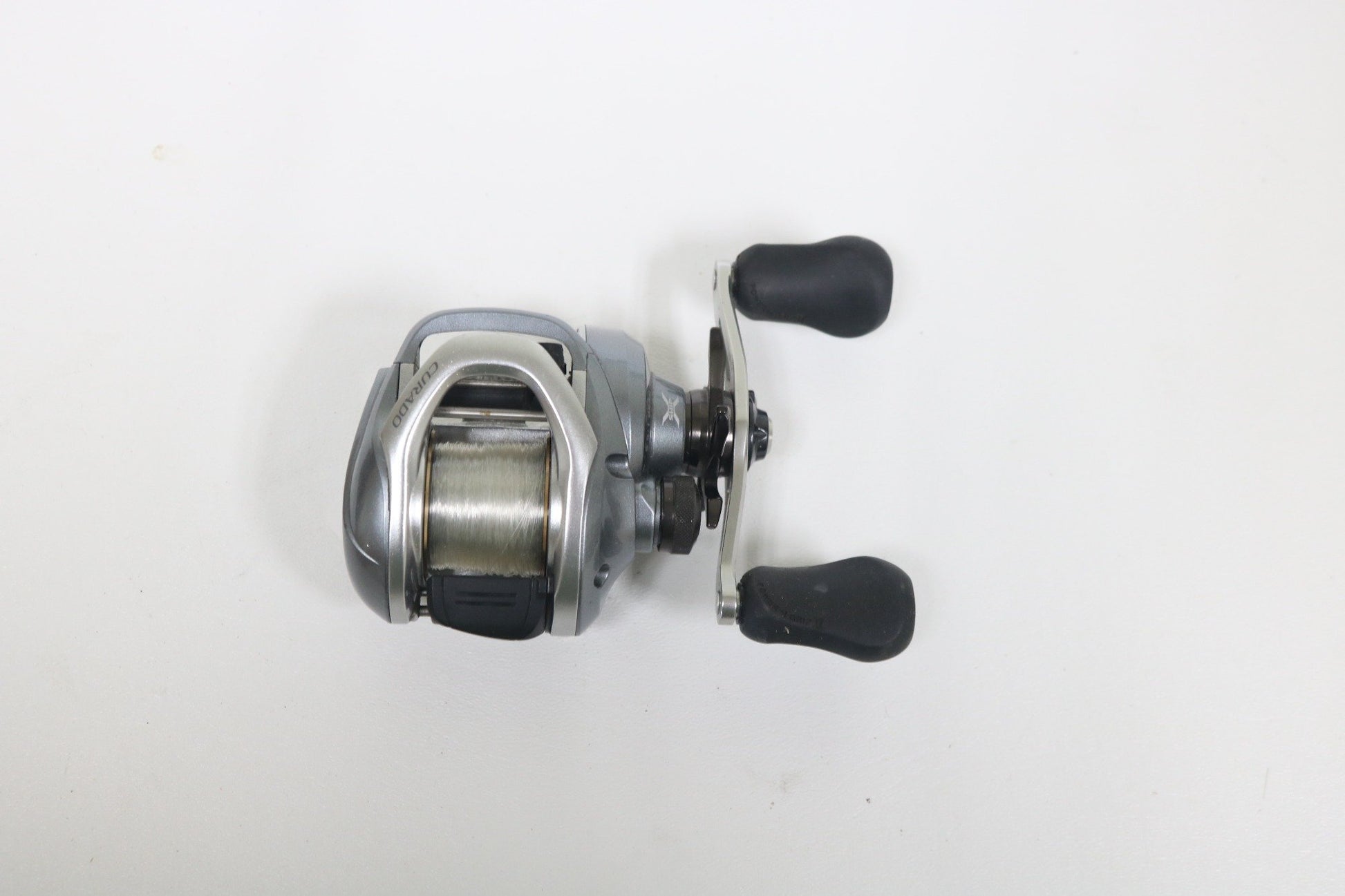 The Shimano Curado I 200PG 5.5:1 Used Casting Reel in excellent condition features a silver design with black handles and spooled line, ideal for anglers upgrading via our Rod and Reel Trade-In or shopping quality Used Gear.