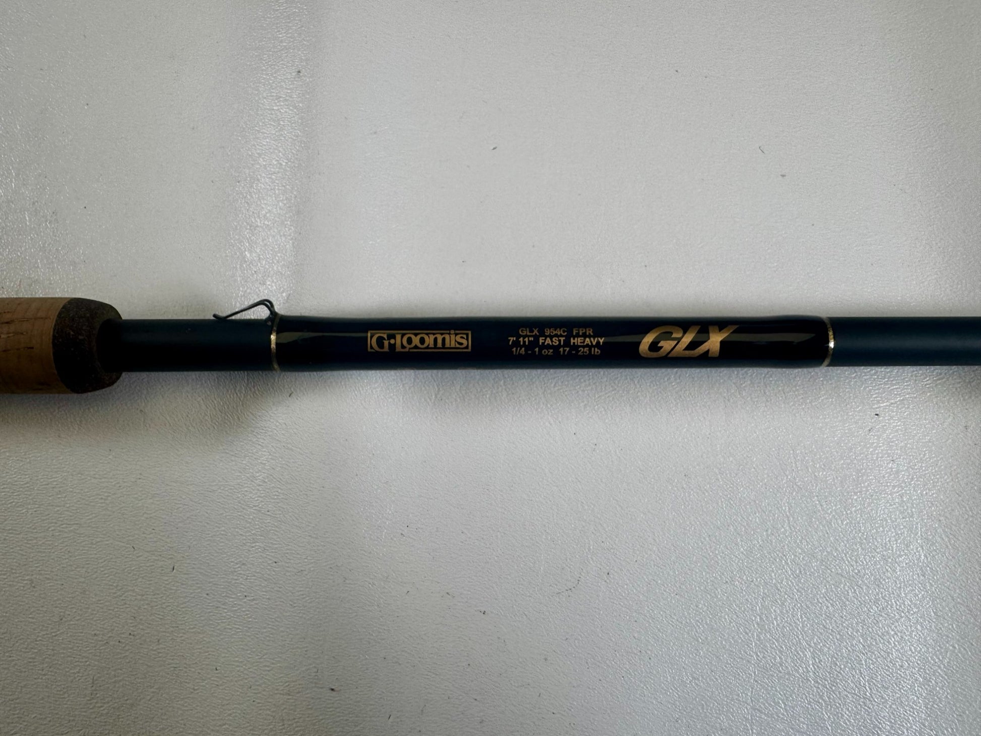 A close-up of the G. Loomis 24 GLX 954c FPR 7'11" Heavy Used Casting Rod in excellent condition, showing gold model details on a dark rod with a cork handle—ideal for those interested in used gear or our Trade-In Program.