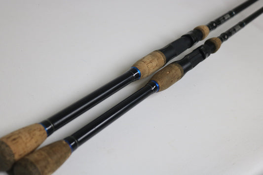 Two SixGill Creature CREC703H and CREC703MH casting rods in good used condition with cork handles lie parallel on a white surface. Trade in your old gear through our Rod and Reel Trade-In Program to earn store credit.