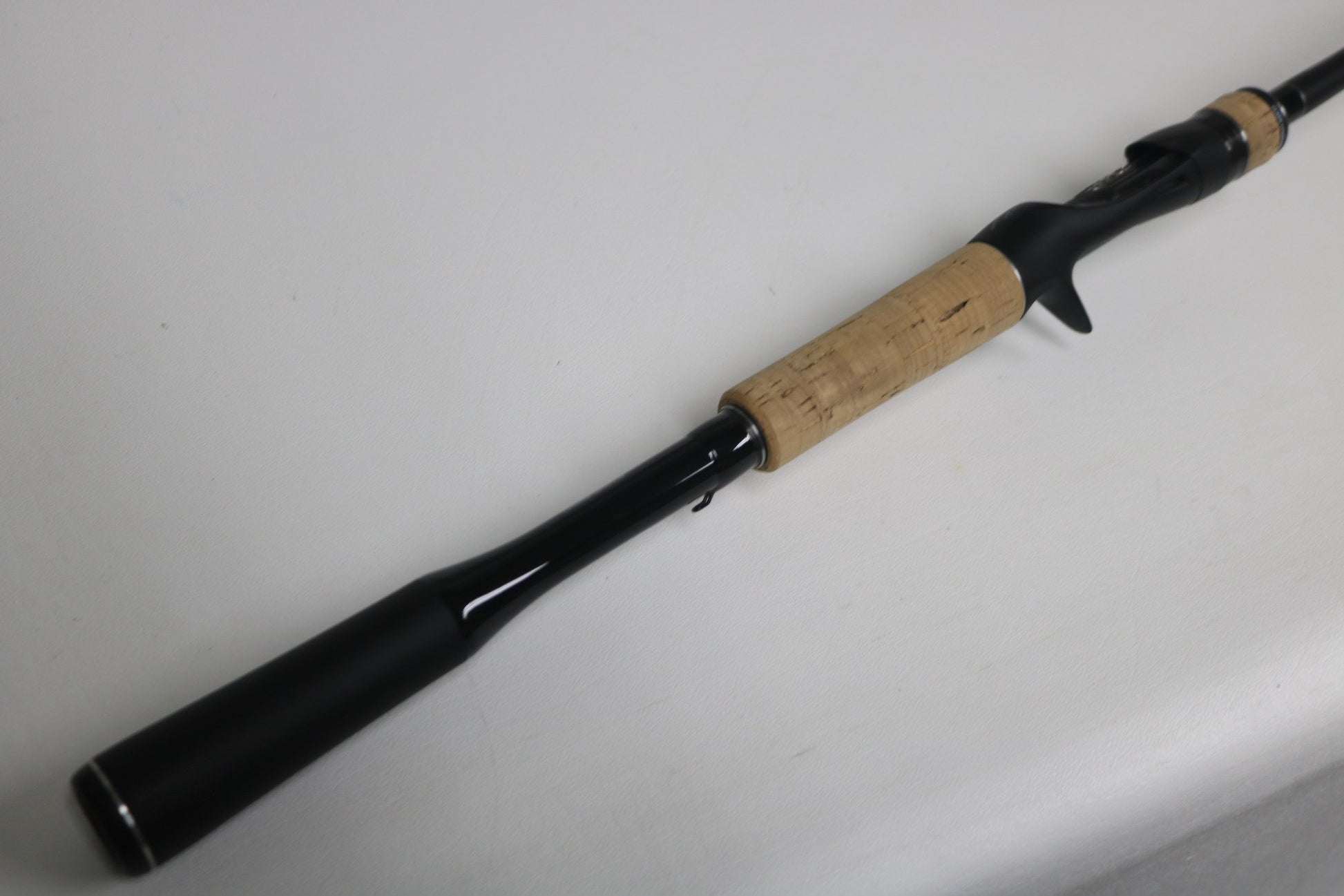 A Shimano Expride B EXC70MGB used casting rod in excellent condition, featuring a black finish and cork handle, is displayed diagonally on a white surface.