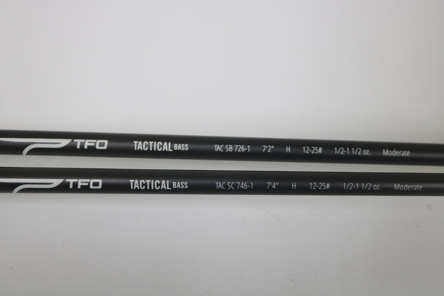Two black Temple Fork TFO Tactical Bass TAC SB 726-1 and TAC SC 746-1 casting rods (used, good condition) are side by side on a white surface with white specs. Trade in used gear for store credit through our Rod and Reel Trade-In Program!.