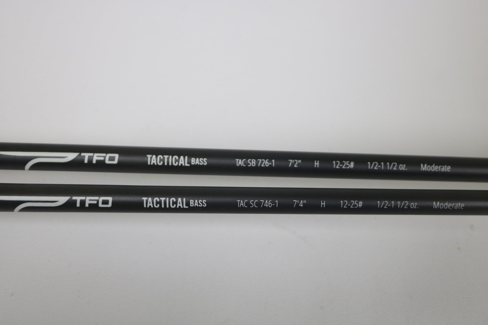 Two black Temple Fork TFO Tactical Bass TAC SB 726-1 and TAC SC 746-1 casting rods (used, good condition) are side by side on a white surface with white specs. Trade in used gear for store credit through our Rod and Reel Trade-In Program!.