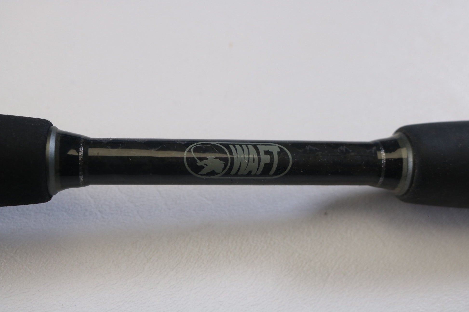 A close-up of the Waft Rods Iron Feather 7'0" Medium Heavy Used Casting Rod handle shows its black, slightly textured grip and white "WAFT" logo with fish graphic—great for trade-ins or checking used gear condition. Brand: Waft.