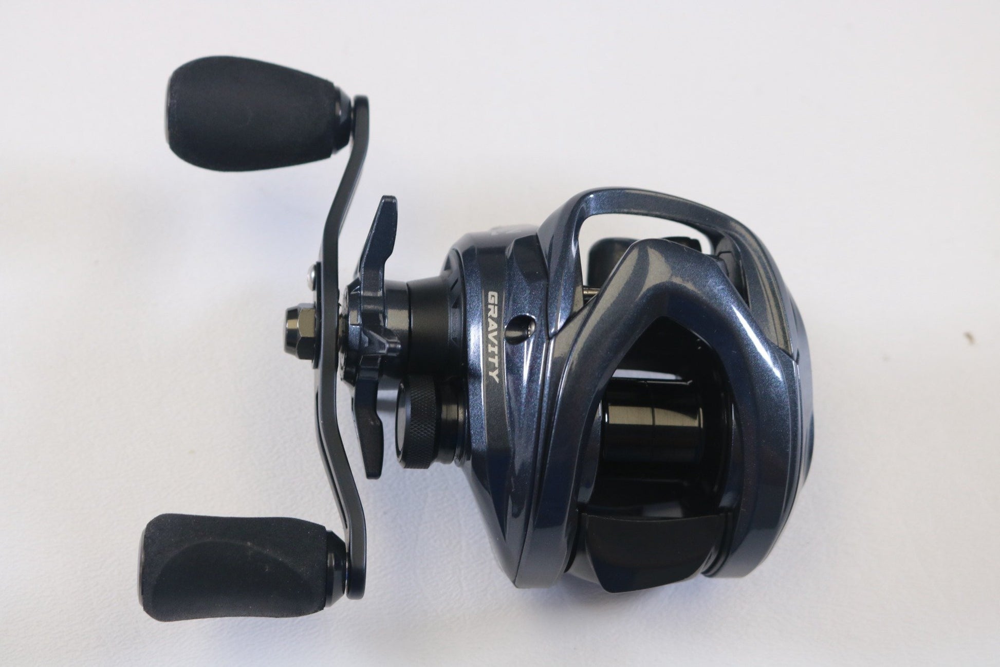 A close-up photo of an Ark Fishing Gravity 3 G3107L 7.1:1 LH Used Casting Reel in excellent condition, dark blue with black handles—ideal for those seeking quality pre-owned gear or eyeing a rod and reel trade-in for store credit.
