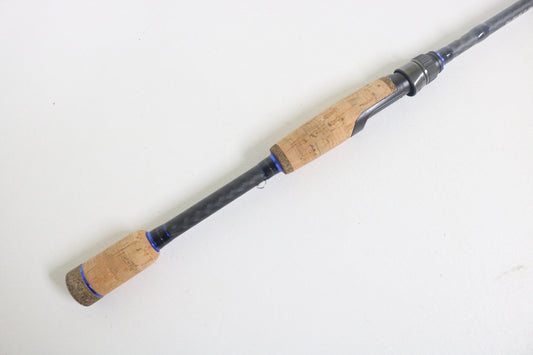 The Dobyns Champion XP DC701SF spinning rod, featuring a carbon fiber shaft with cork handles that are light brown with darker speckles, is resting on a white surface. The rod's components are connected by blue accents and it is in excellent condition.