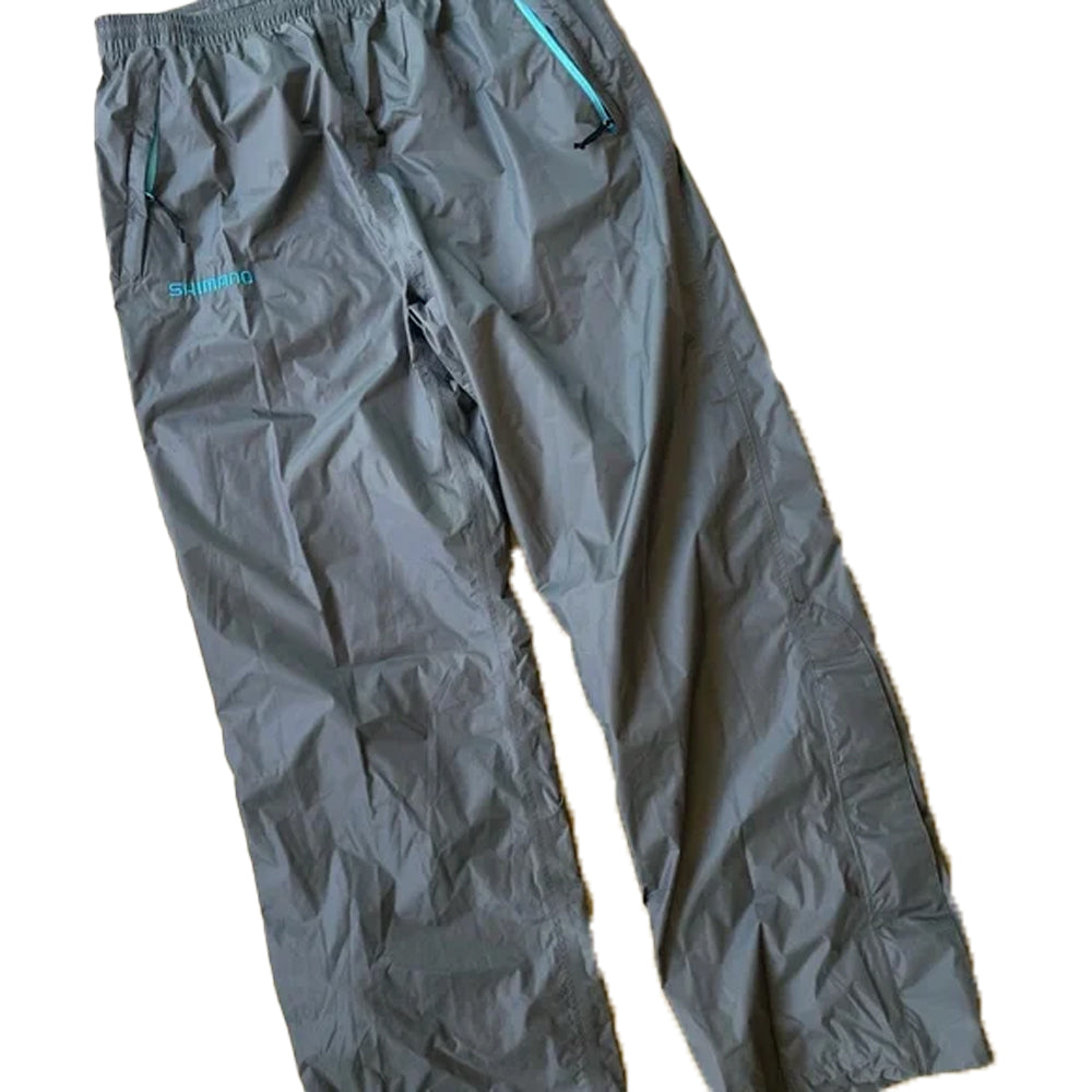 Shimano Lightweight Rain Pants in charcoal, size XXL, feature an elastic waistband and blue zipper pockets. The "SHIMANO" brand is visible on the left pocket. Shown laid flat on a white background.